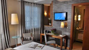 Quarto de hotel com cama, escrivaninha, cadeira, televisão e banheiro visível através de uma porta aberta.