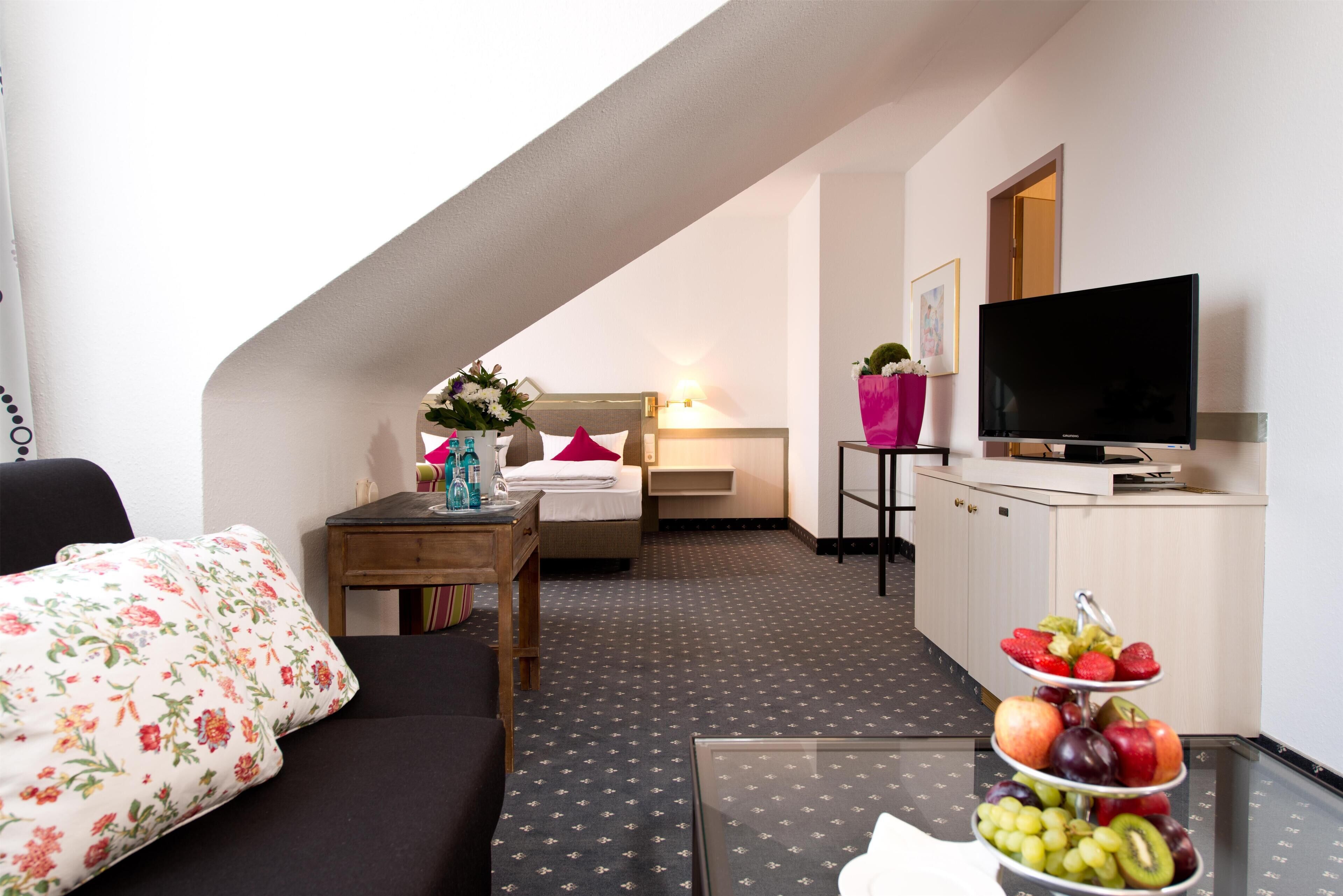 A hotel room with a bed, a TV, a small table with a vase of flowers, and a fruit tray.