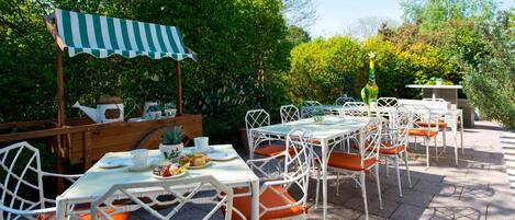 An outdoor patio with white metal chairs and tables, a striped awning, and lush greenery in the background.