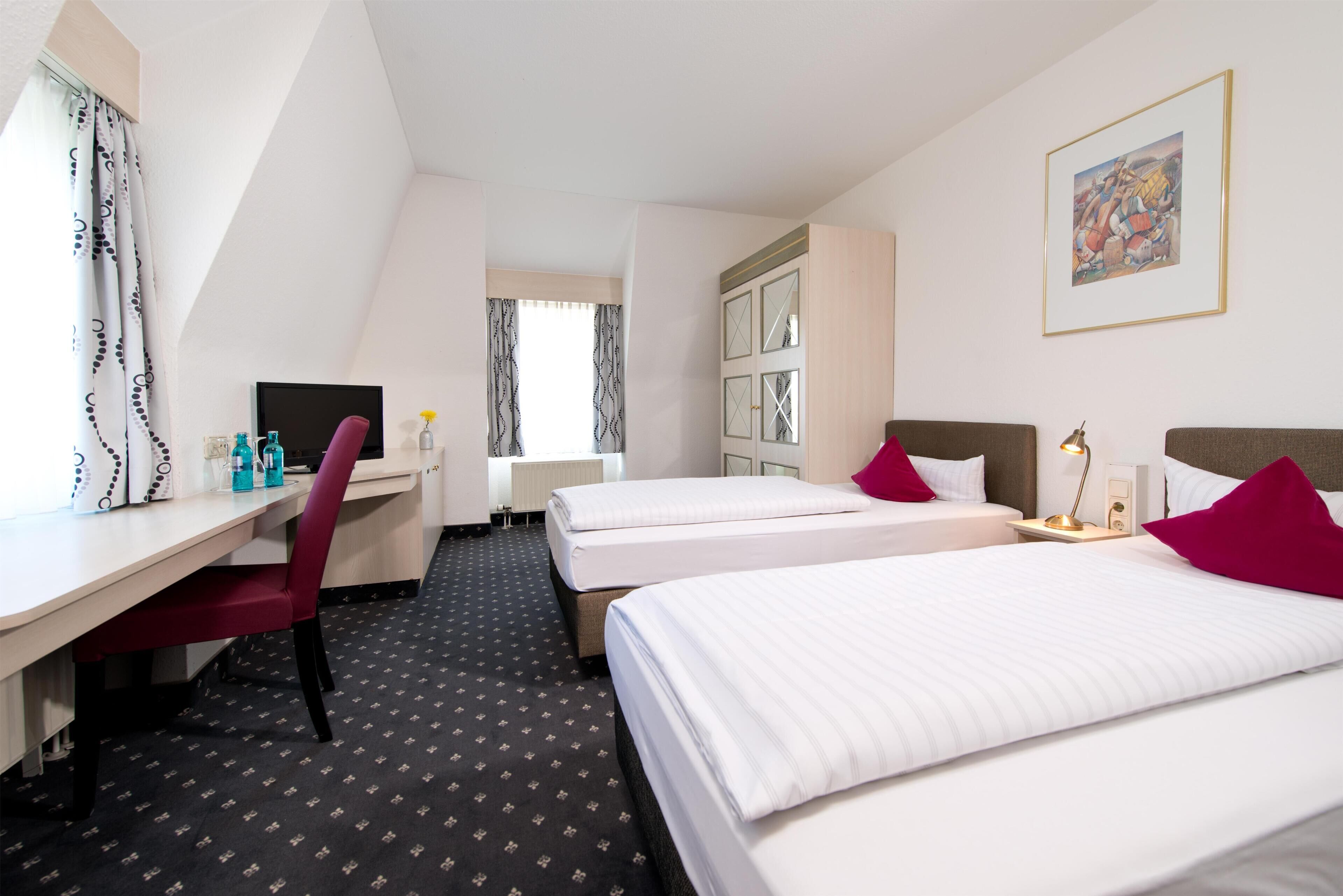 Habitación de hotel con dos camas, un escritorio, una silla y una ventana con cortinas.