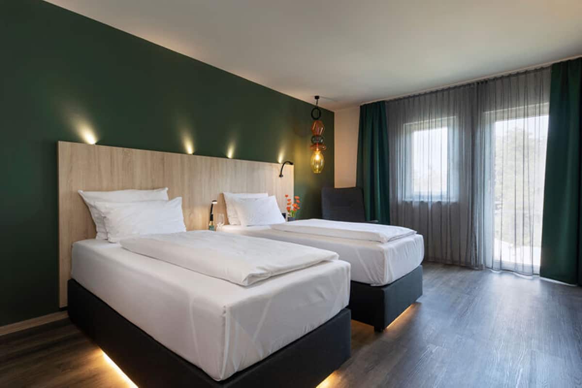 A hotel room with two beds, a wooden headboard, a wall-mounted light fixture, and a large window with curtains.