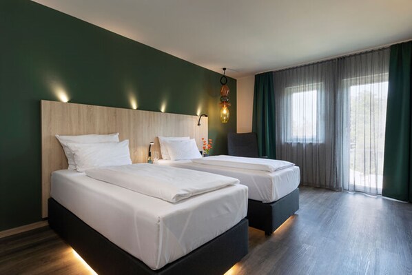 Habitación de hotel con dos camas, cabecero de madera, lámpara de pared y una ventana grande con cortinas.