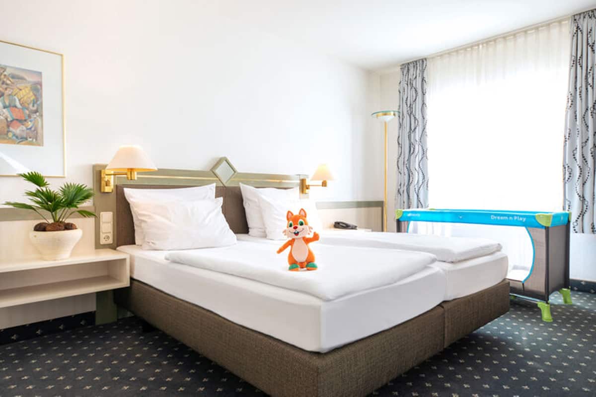 A hotel room with a bed, a nightstand with a lamp, a desk, a chair, a window with curtains, and a small plant.