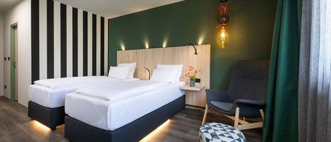 Una habitación de hotel moderna con dos camas, un cabecero de madera, una mesita de noche con lámpara, un sillón gris y un reposapiés estampado.