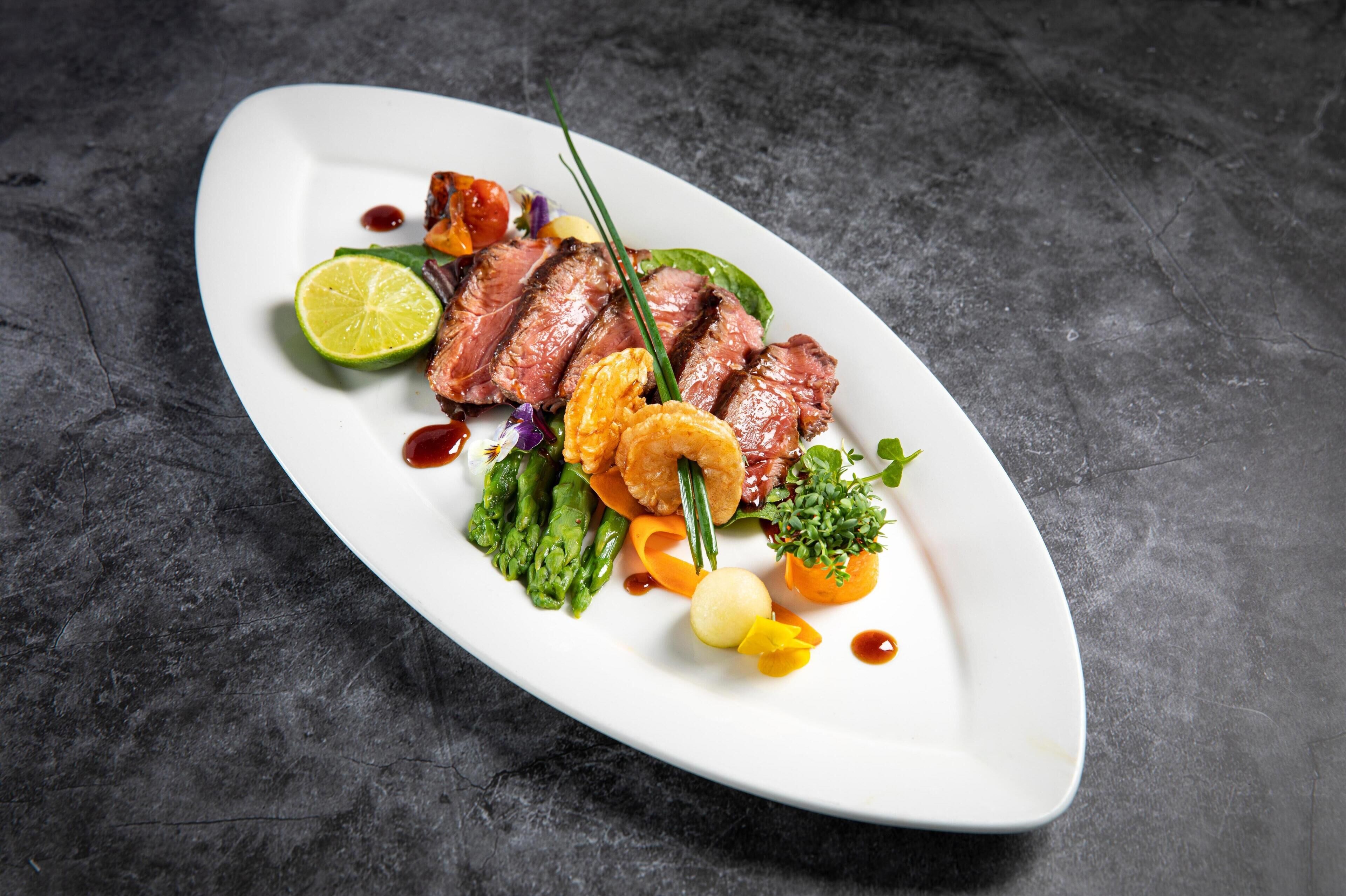 A white plate with steak, asparagus, and vegetables on a dark surface.