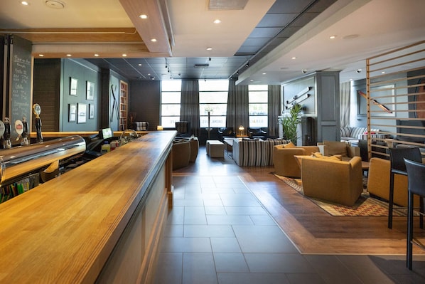 A modern bar area with a wooden counter, seating with cushions, and large windows providing natural light. - Scandic Helsinki Aviacongress (Vantaa)
