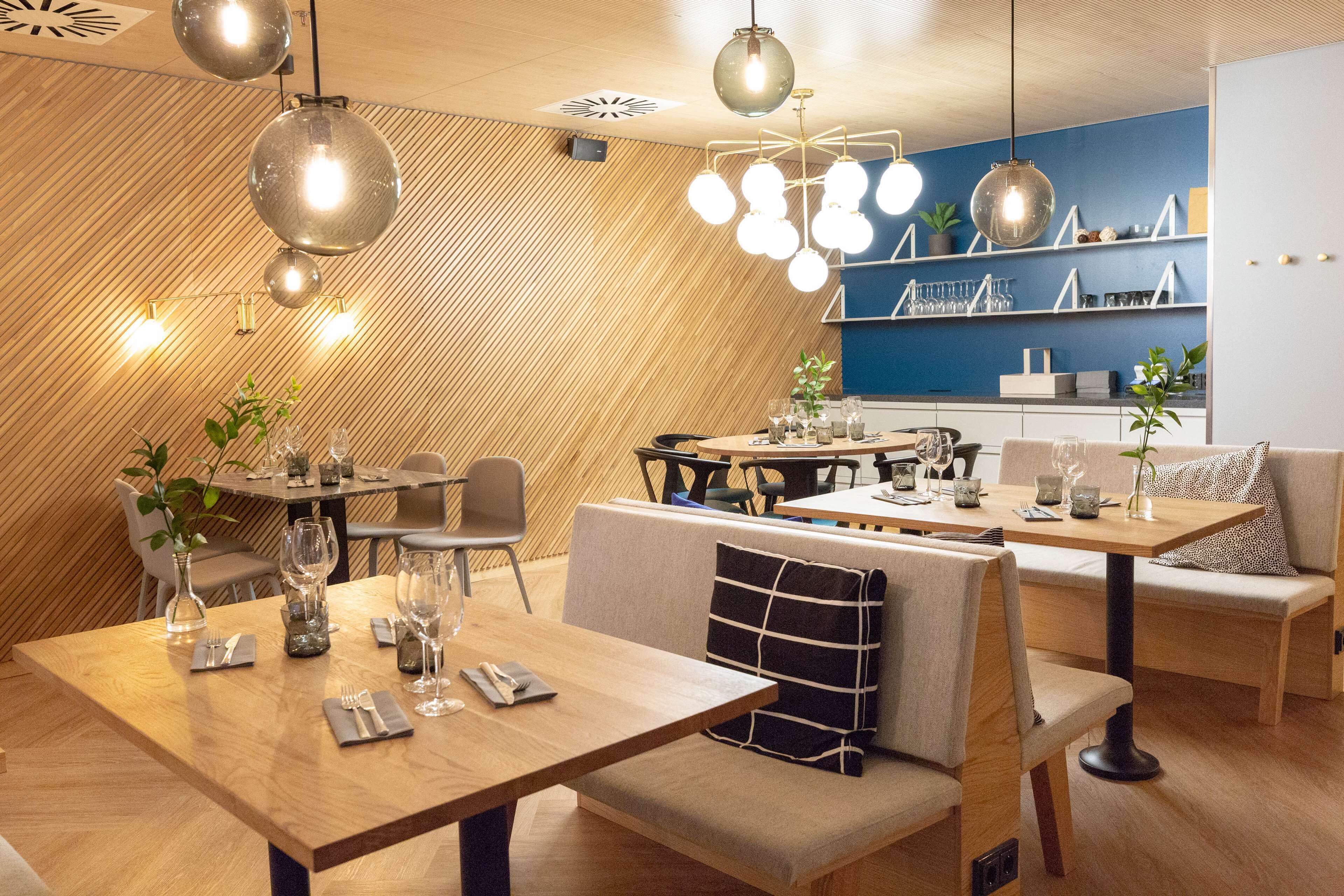 Um restaurante moderno com mesas e cadeiras de madeira, luminárias pendentes e uma parede azul como detalhe.