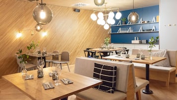 Um restaurante moderno com mesas e cadeiras de madeira, luminárias pendentes e uma parede azul como detalhe.