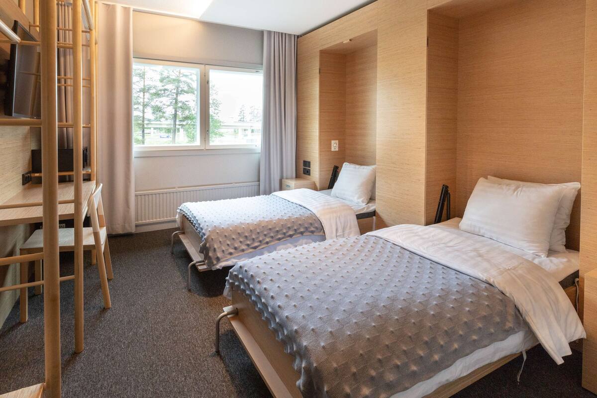 A hotel room with two beds, a wooden wardrobe, a desk, and a window with curtains.