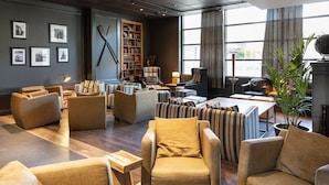 A modern lounge area with multiple seating options, including sofas and armchairs, a bookshelf, and a large window providing natural light. - Scandic Helsinki Aviacongress (Vantaa)
