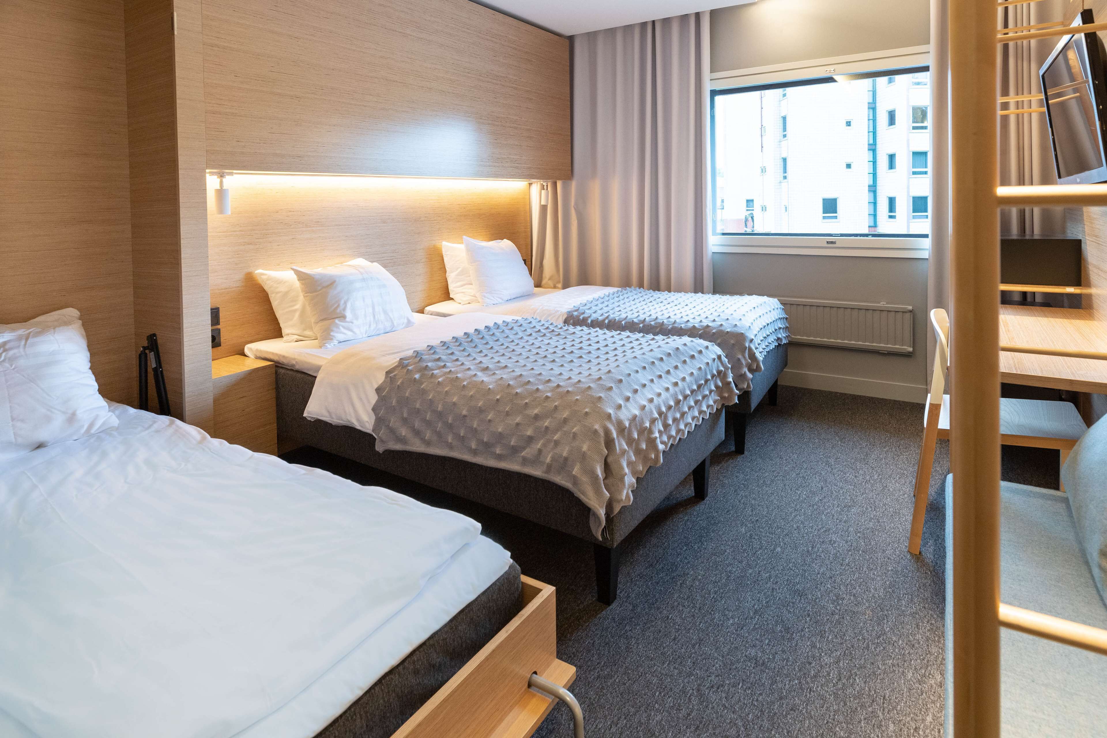 Ein Hotelzimmer mit zwei Betten, einem Schreibtisch und einem Fenster mit Blick auf Gebäude.