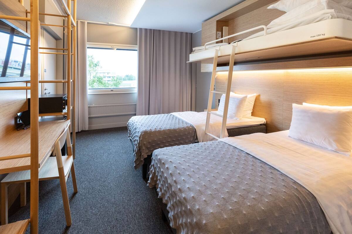 A compact hotel room with bunk beds, a desk, and a window with curtains.
