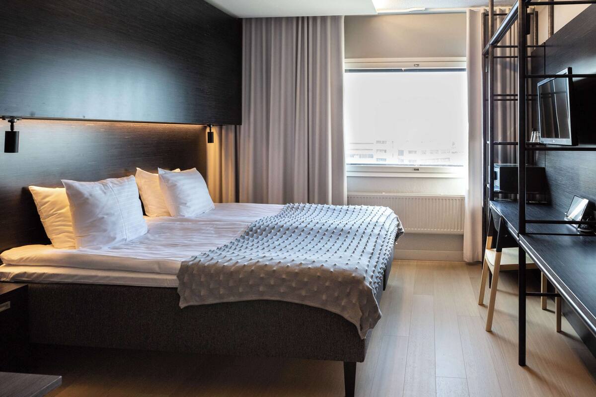A modern hotel room with a large bed, a desk, and a view of the city through the window.