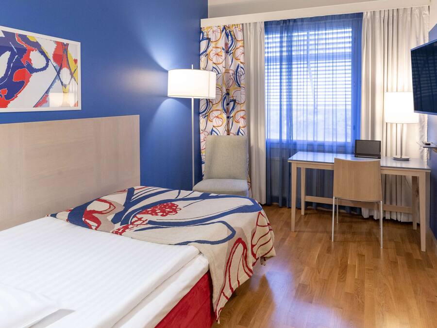 Quarto de hotel com cama, escrivaninha, cadeira, televisão e janela com cortinas.