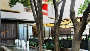 Un patio con fuente de agua, árboles y un edificio de fachada roja y blanca con grandes ventanales.