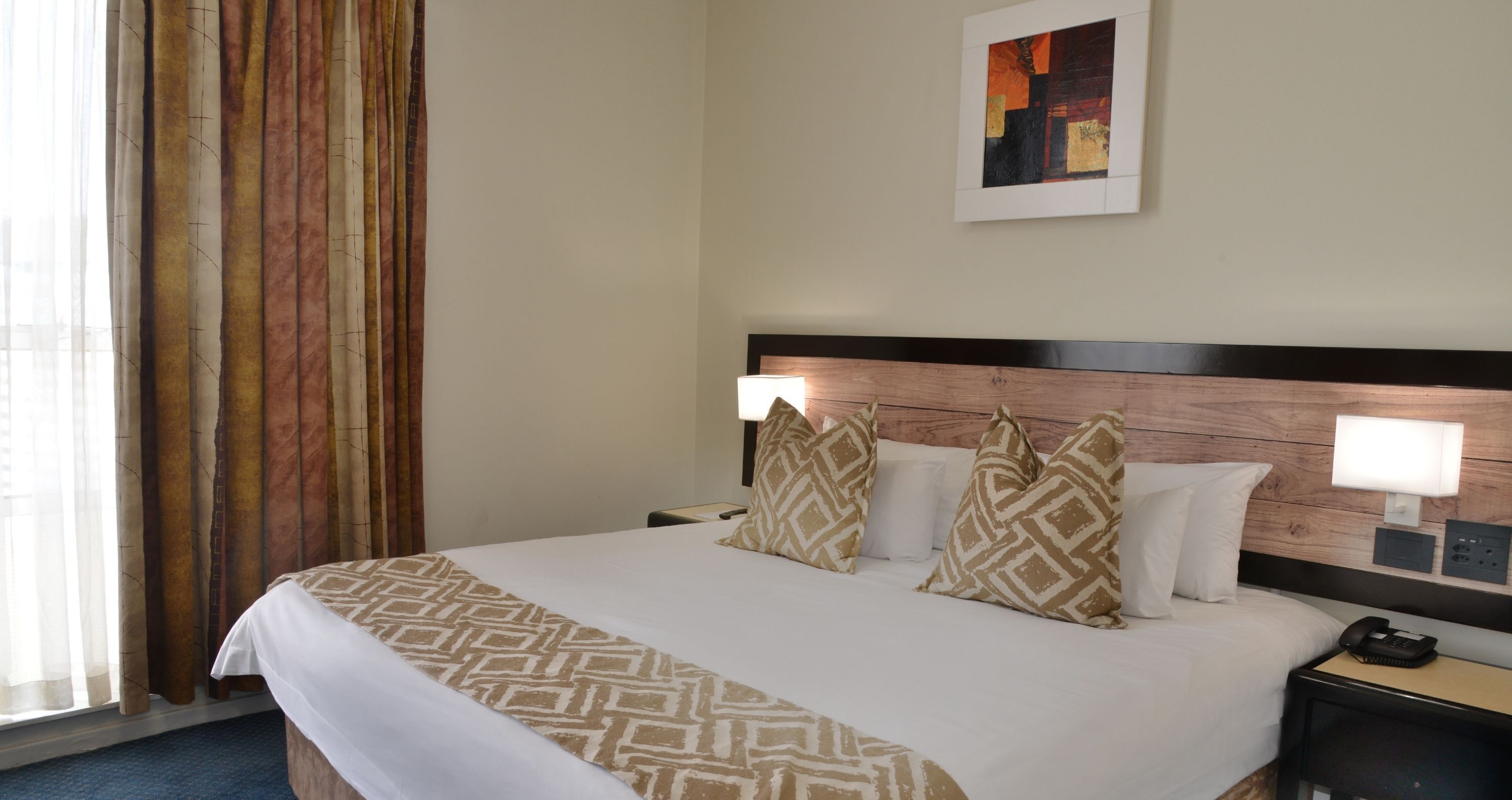 A hotel room with a large bed, two bedside lamps, a nightstand with a phone, and a painting on the wall.
