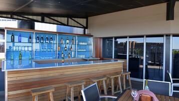 Uma área moderna de bar ao ar livre com bancos de madeira, balcão de bar com tampo de vidro e prateleiras cheias de garrafas e copos.