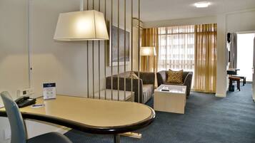 A hotel room with a desk, two armchairs, a sofa, a coffee table, and a large window with curtains.