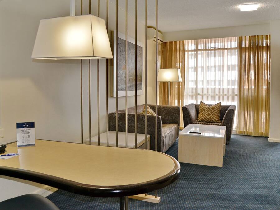A hotel room with a desk, two armchairs, a sofa, a coffee table, and a large window with curtains.