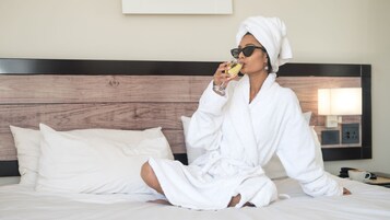 Una persona sentada en una cama con bata blanca, ropa de cama blanca, cabecera de madera y una mesita de noche con una lámpara y una taza.