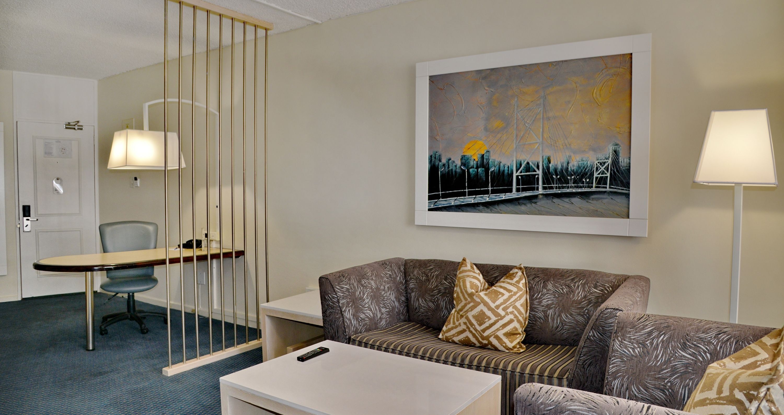 A hotel room with a sofa, a small table, a chair, a desk, and a painting on the wall.