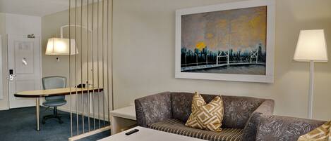 A hotel room with a sofa, a small table, a chair, a desk, and a painting on the wall.