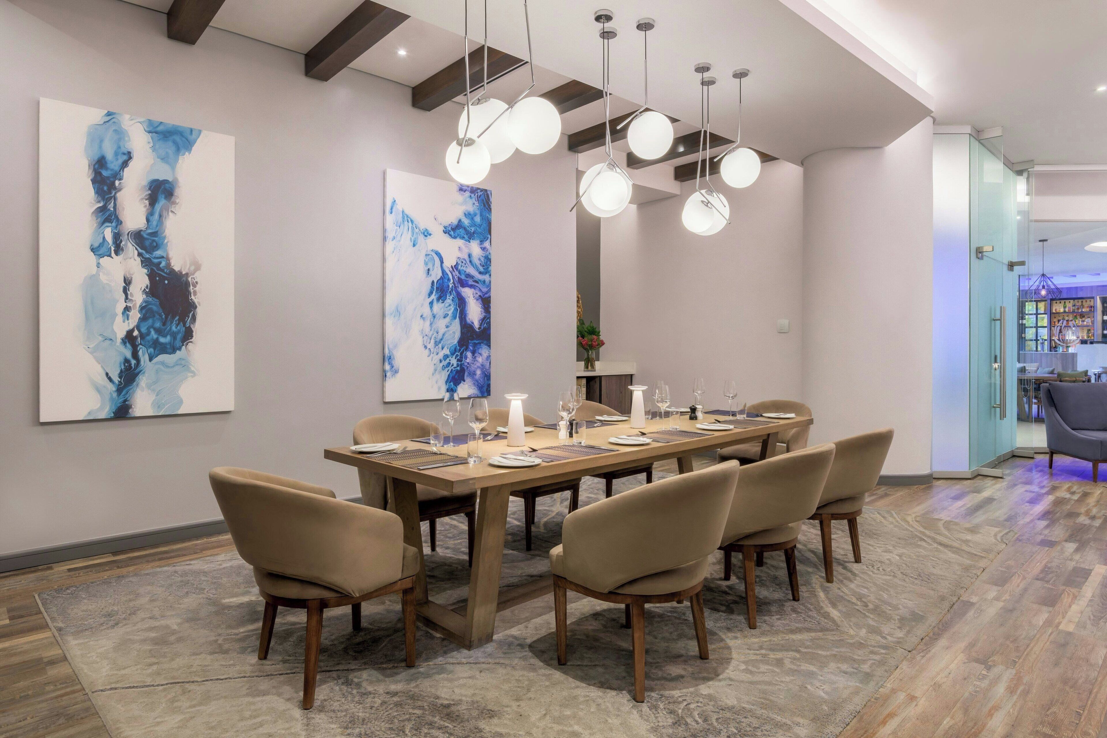 Un espace repas moderne avec une longue table en bois, dressée pour six personnes, entourée de six chaises. Les murs sont ornés de peintures abstraites, un lustre est suspendu au-dessus de la table et une cloison en verre se trouve à l’arrière-plan.
