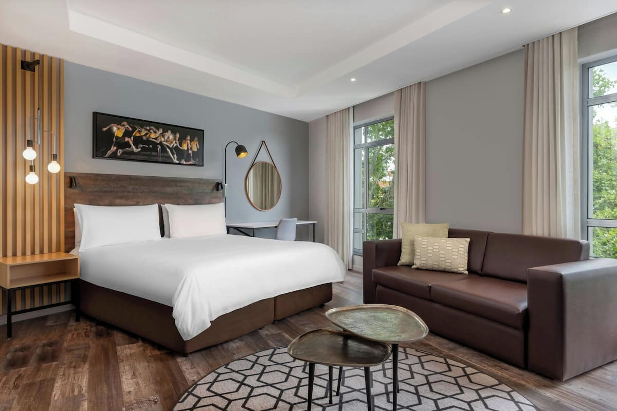 Une chambre d’hôtel moderne avec un grand lit, un canapé marron, une petite table et une grande fenêtre.