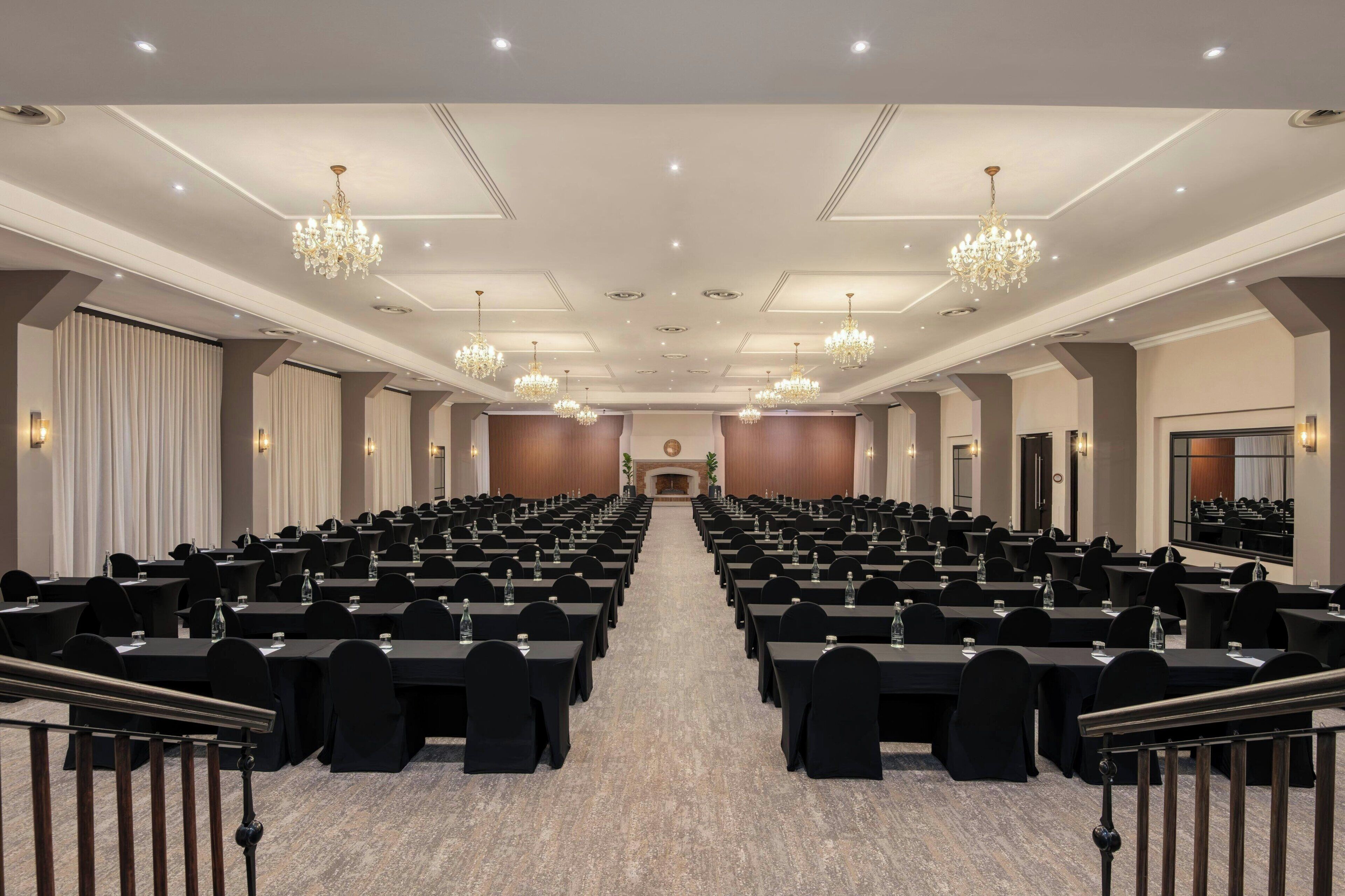Un salón de conferencias con filas de mesas y sillas, candelabros y un escenario al frente.