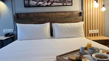 A hotel room with a bed, white pillows, a painting on the wall, a bedside table with a lamp, and a tray with breakfast items.
