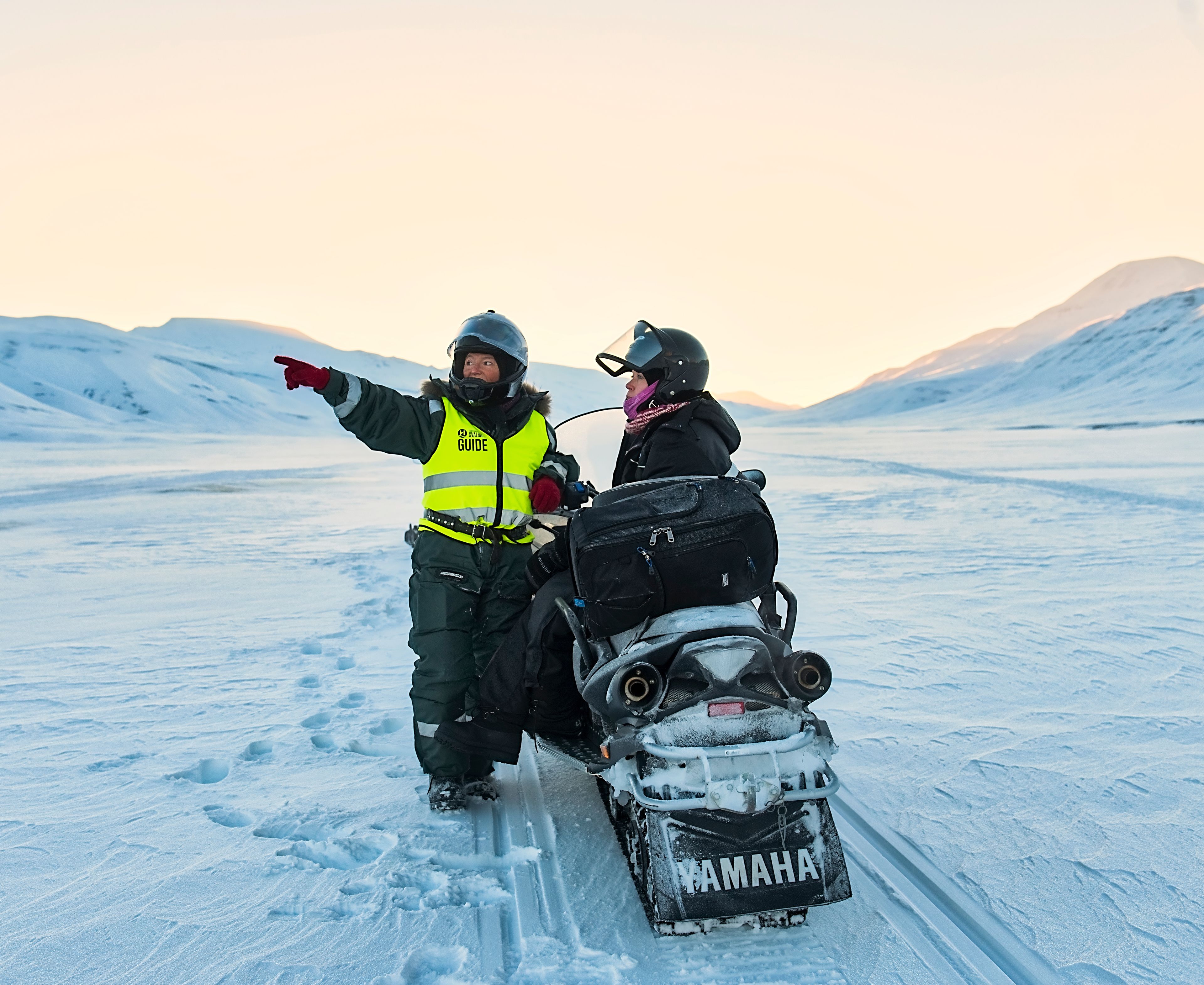 A snowmobile with a Yamaha logo on the front, a person pointing towards the horizon, and a guide in a high-visibility vest.