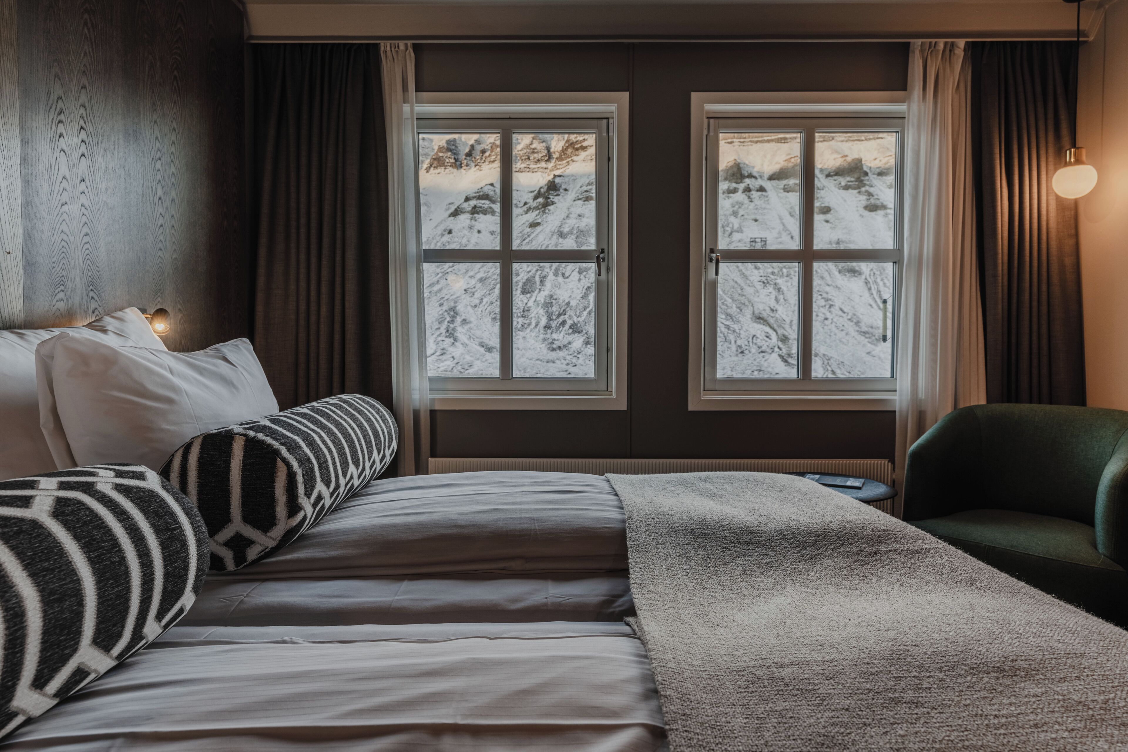 A hotel room with a bed, two windows with a mountain view, a green armchair, and a wall-mounted lamp.