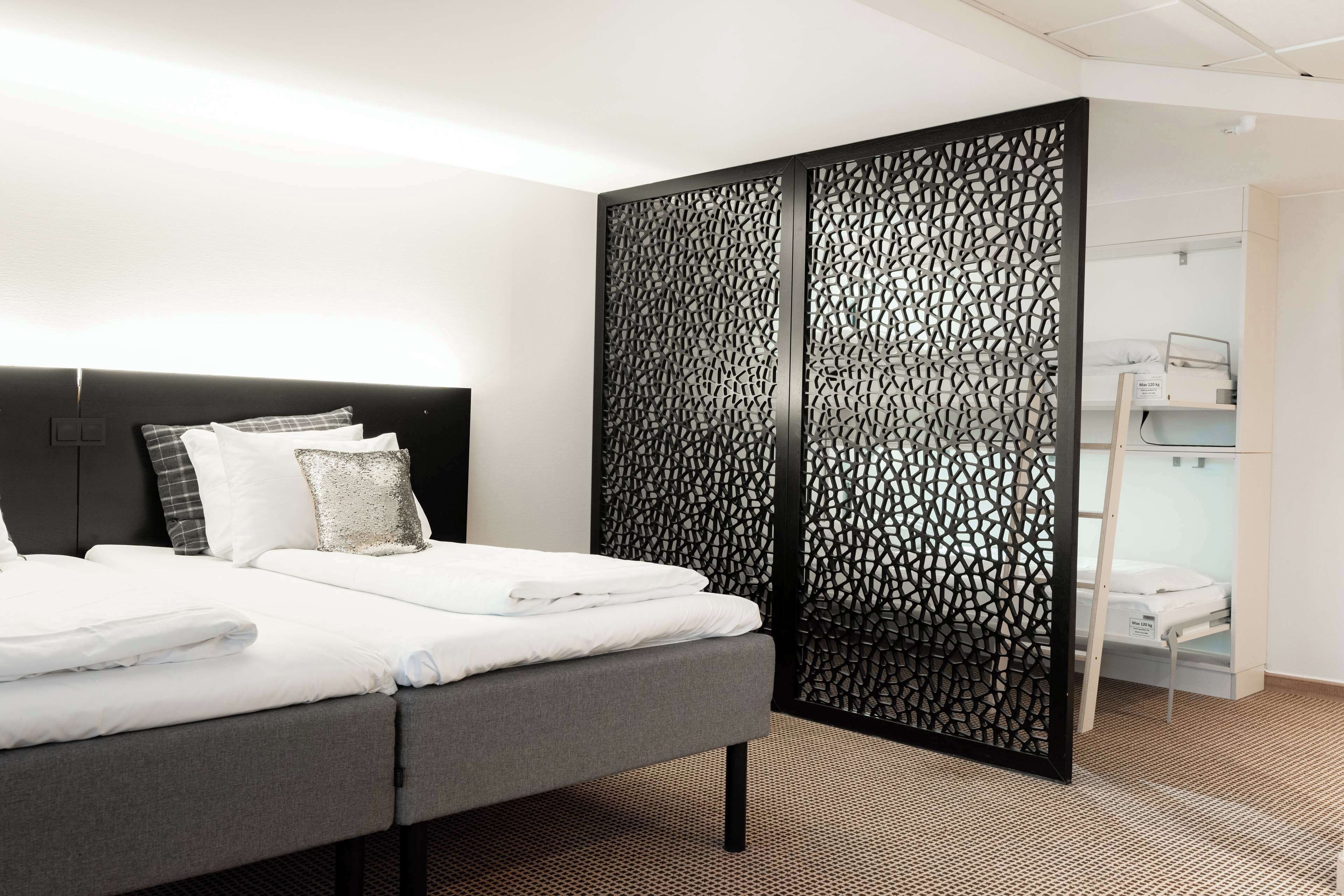 Una habitación de hotel moderna con una cama grande, un panel decorativo negro y una mesita de noche pequeña con una lámpara.
