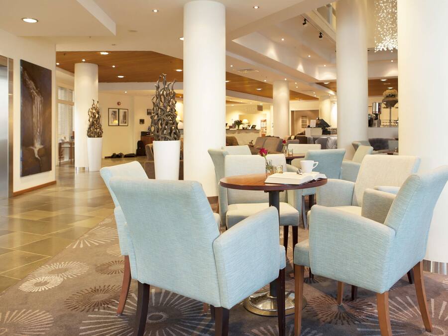 A spacious lobby with light blue upholstered chairs, a round wooden table, and a decorative metal sculpture.