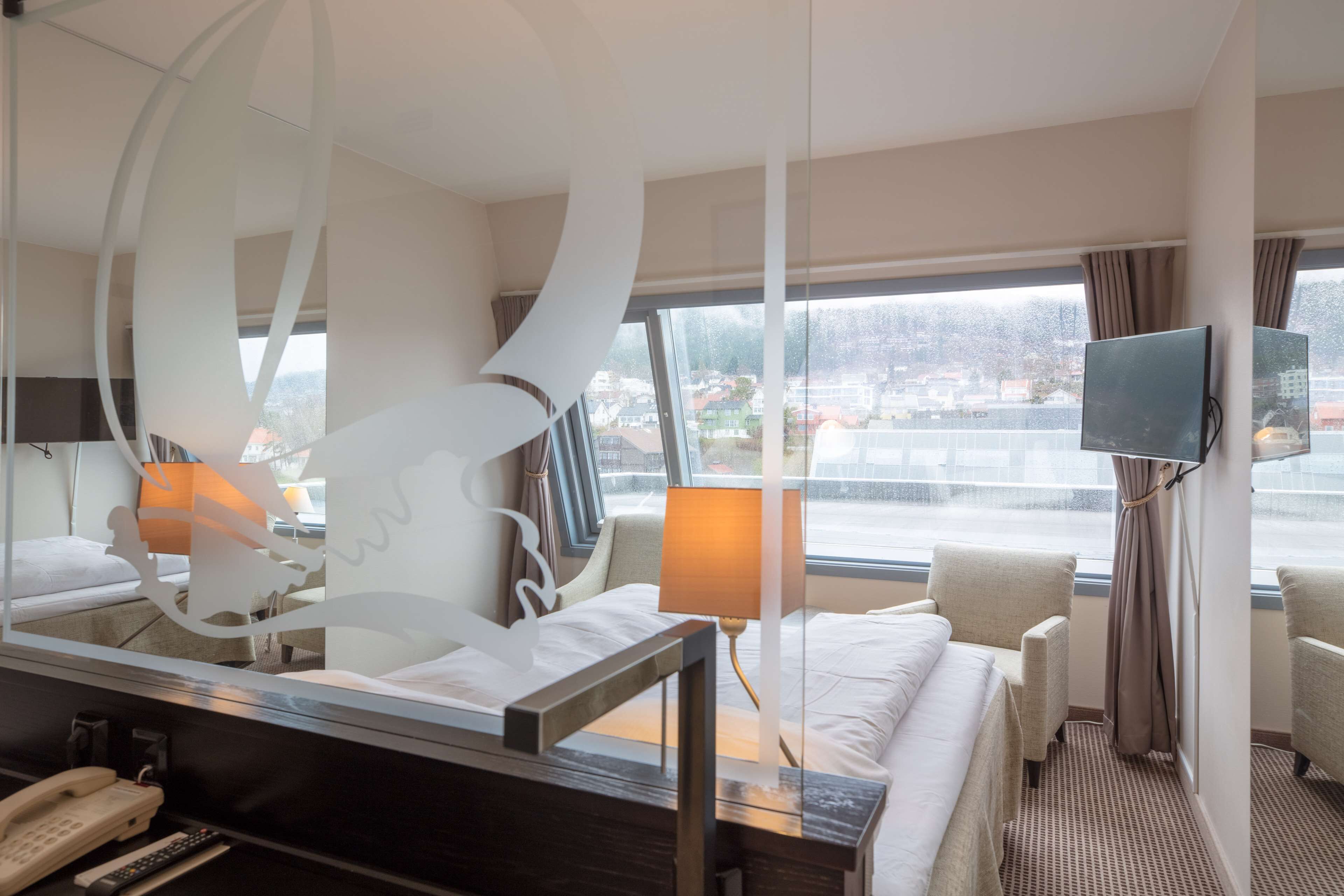 A hotel room with a bed, a desk with a telephone and remote control, a TV mounted on the wall, and a window with a view of buildings and a snowy landscape.