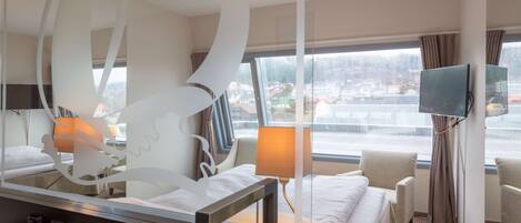 Habitación de hotel con cama, escritorio con teléfono y control remoto, televisor montado en la pared y ventana con vistas a edificios y un paisaje nevado.