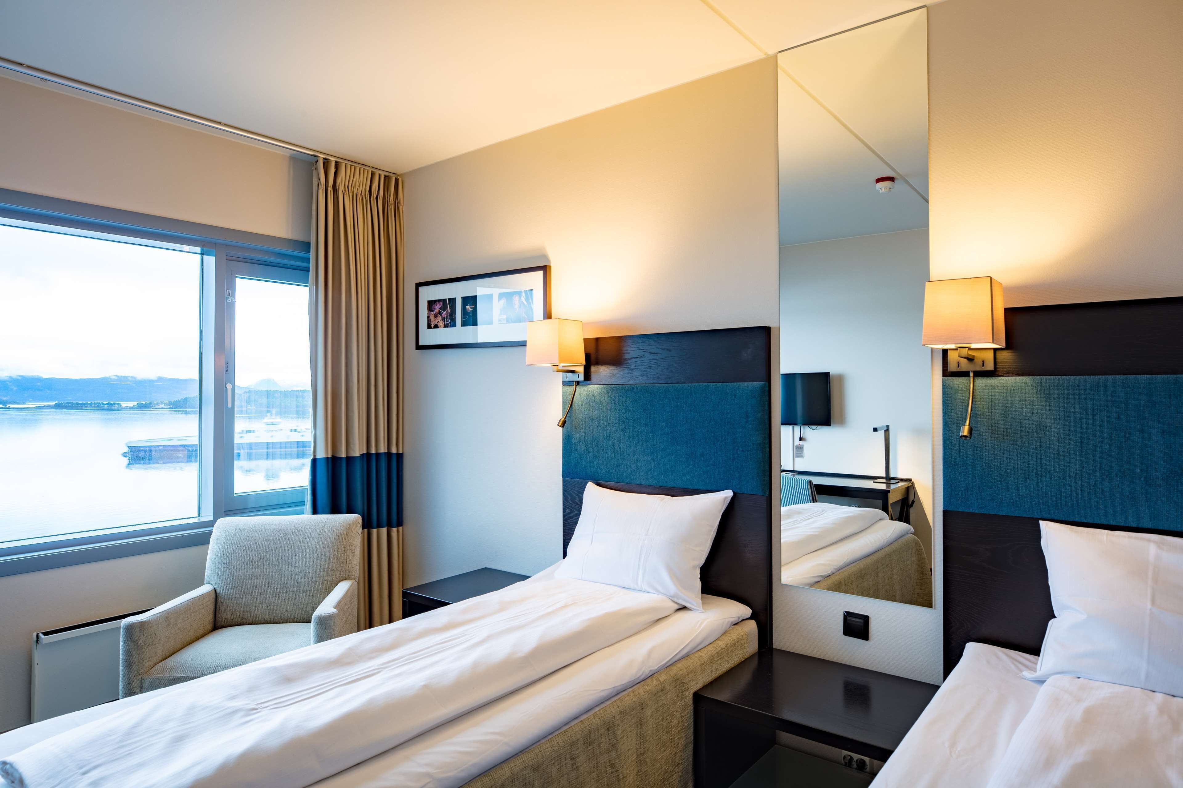 Een hotelkamer met een bed, een stoel, een bureau, een lamp, een spiegel en een raam met gordijnen.