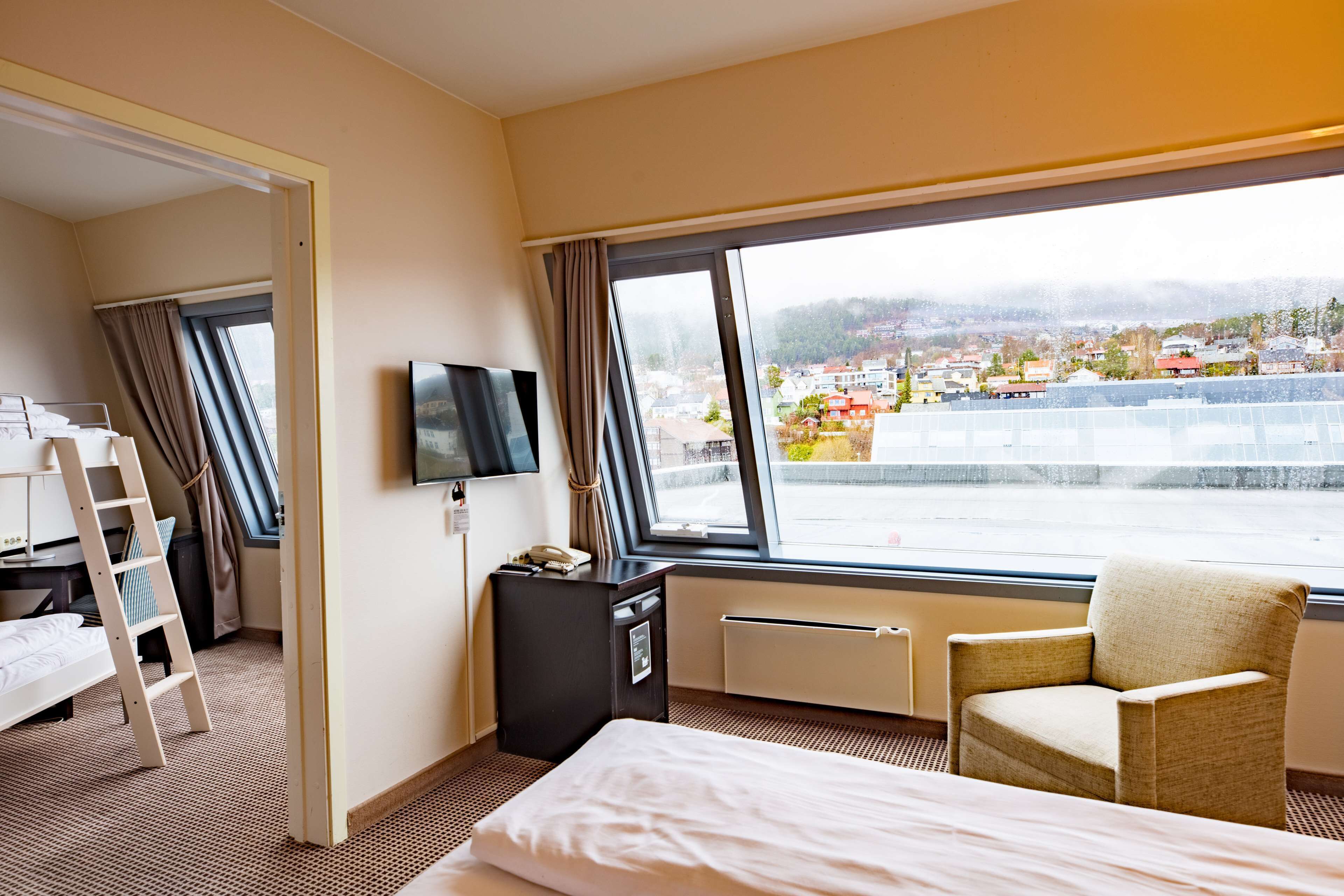 A hotel room with a bed, a desk, a chair, a TV, and a large window offering a city view.