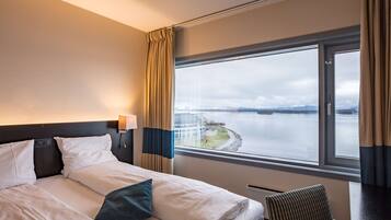 Habitación de hotel con una cama, una silla, una ventana con vista a un cuerpo de agua y un puente, y una lámpara en la pared.