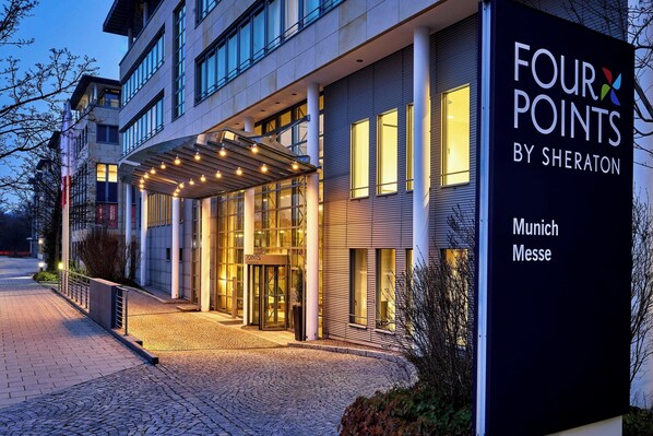 Een hotelingang met een overdekte doorgang, glazen deuren en een bord waarop 'FOUR POINTS BY SHERATON Munich Messe' staat.