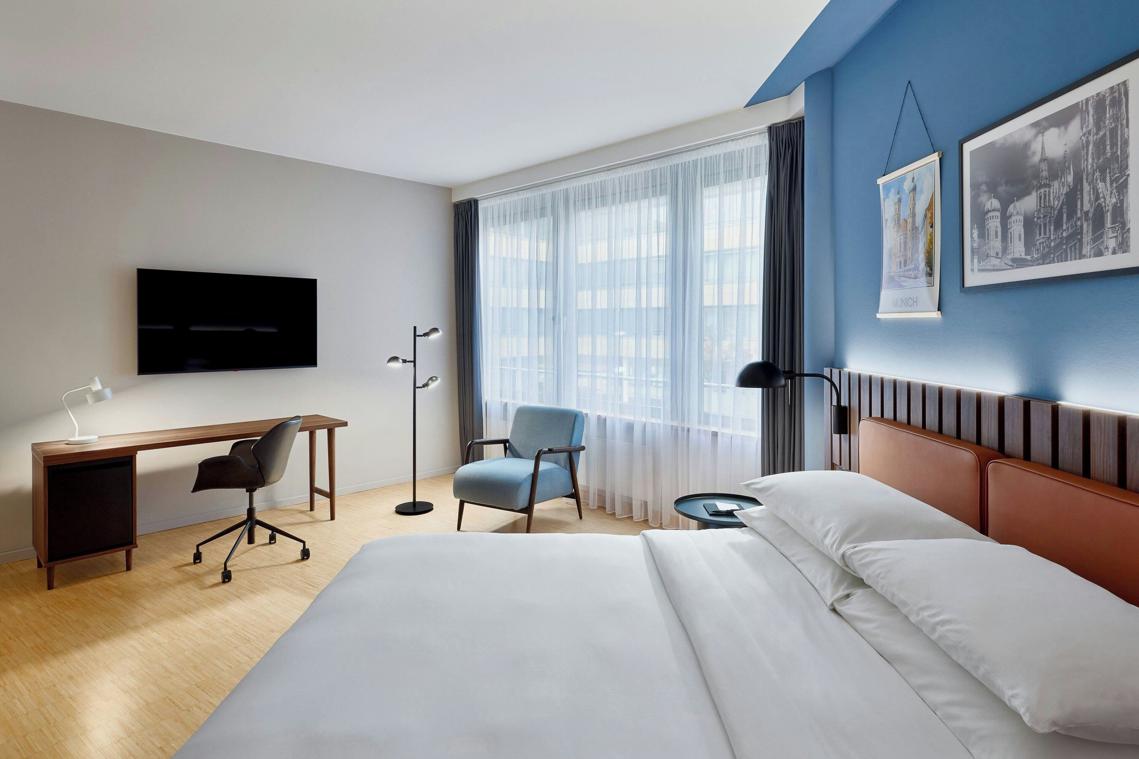 A hotel room with a bed, a desk, a chair, a TV, and a window with curtains.