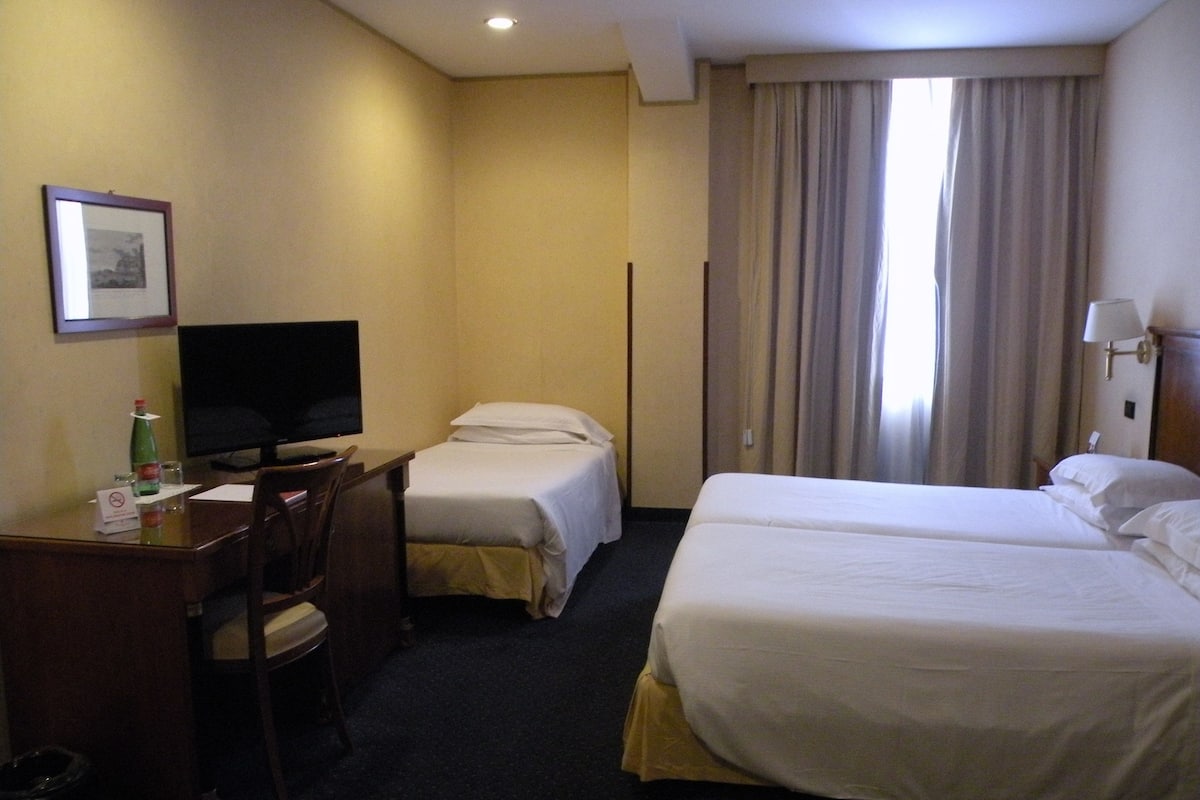 Une chambre d’hôtel avec deux lits simples, un bureau avec une chaise, une télévision et une fenêtre avec des rideaux.