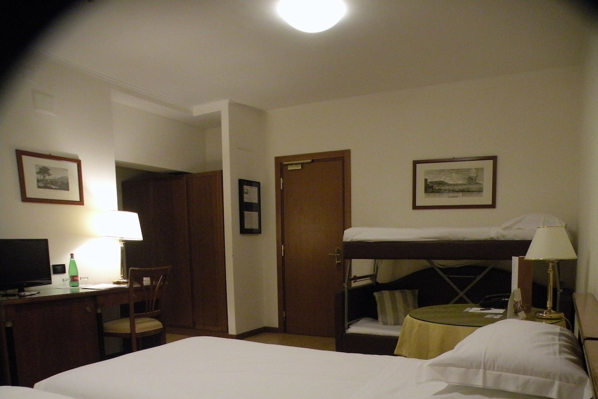 Une chambre d’hôtel avec un lit superposé, un bureau avec une lampe, une chaise et des tableaux encadrés accrochés aux murs.