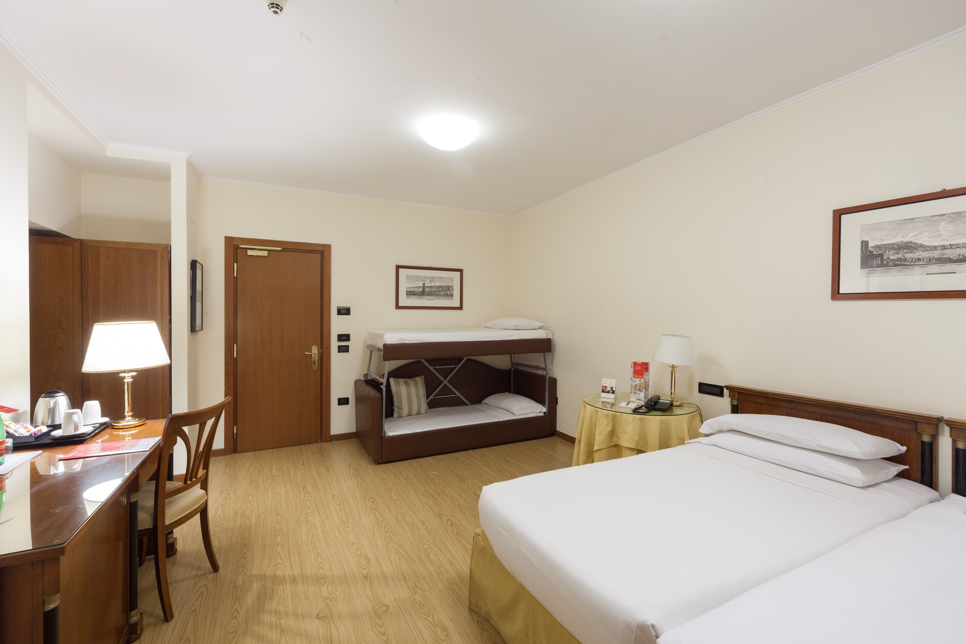 A hotel room with a bunk bed, a desk with a lamp, a chair, a bedside table with a lamp, and a framed picture on the wall.