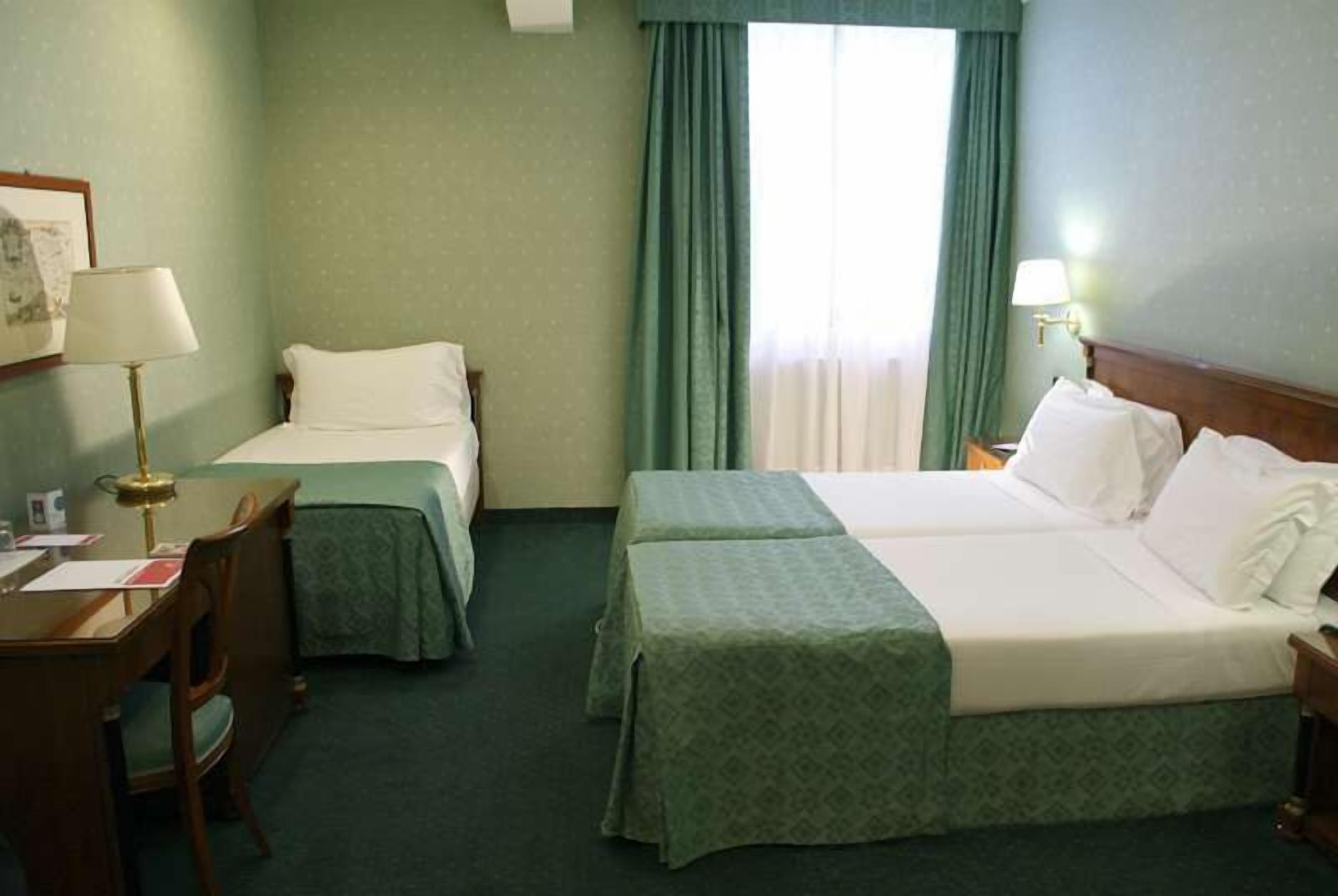 Una camera d'albergo con due letti singoli, una scrivania con lampada e una finestra con tende.