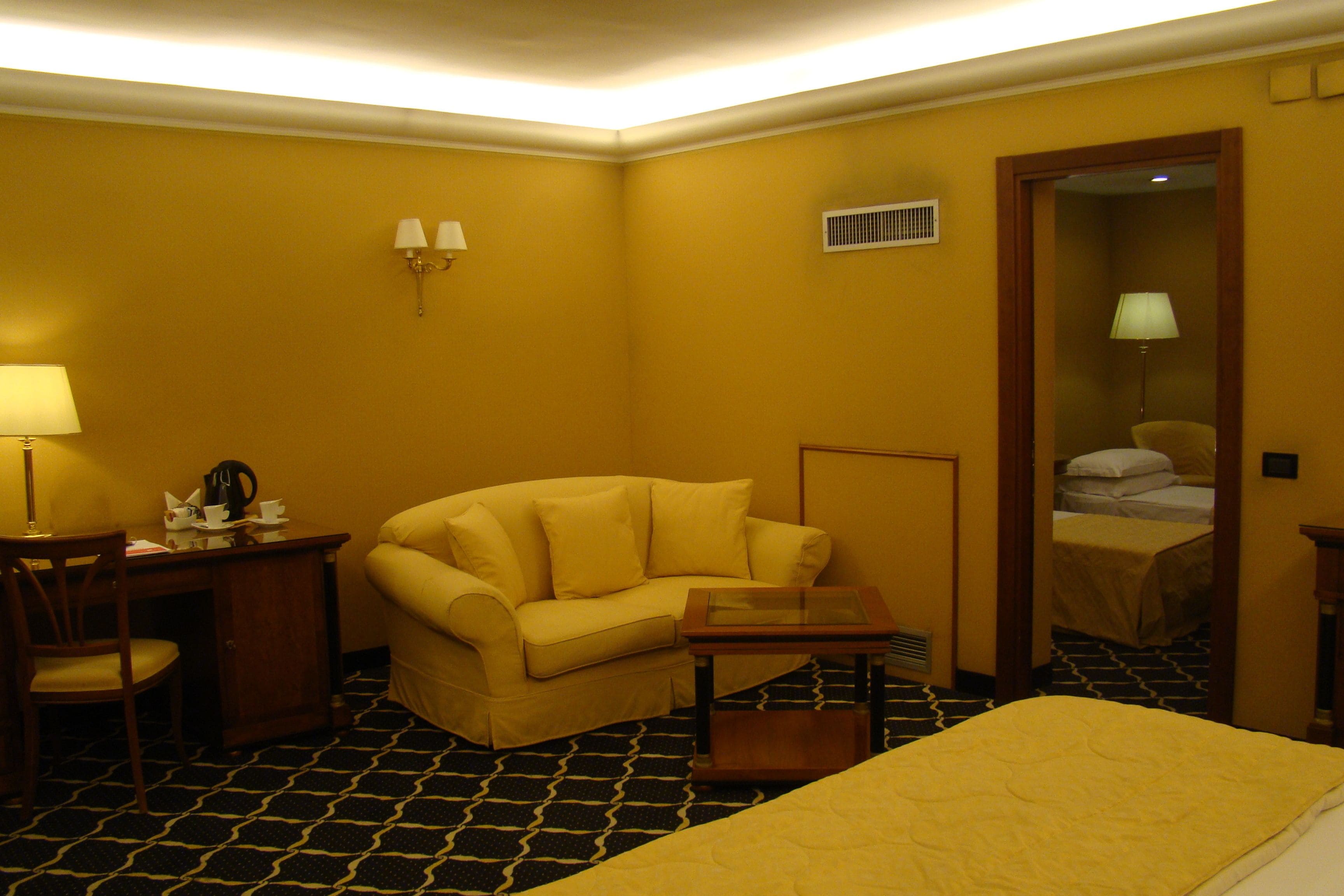 A hotel room with a yellow sofa, a small table, a desk with a lamp, and a bed with a yellow bedspread.