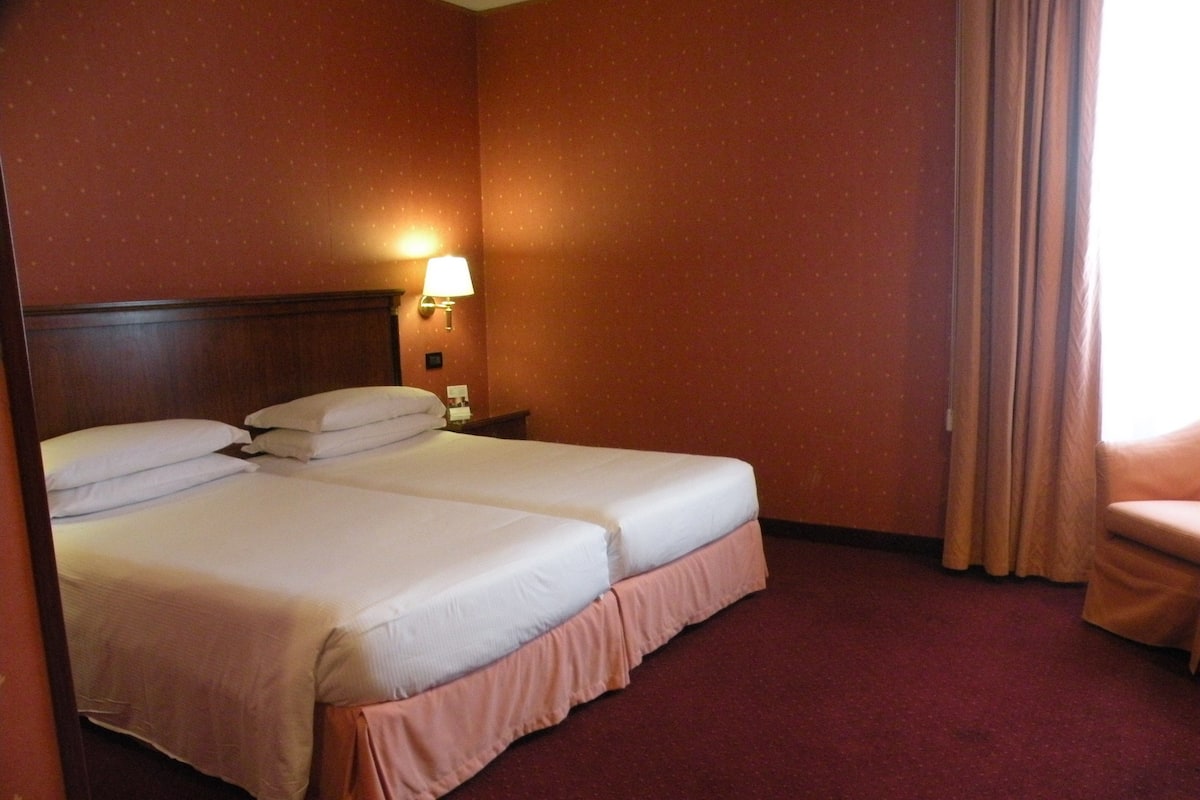 Une chambre d’hôtel avec deux lits, une table de chevet, une lampe et une fenêtre avec des rideaux.