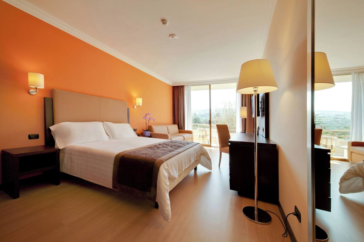 Ein Hotelzimmer mit einem großen Bett, Nachttischlampen, einem Schreibtisch und Blick ins Freie.