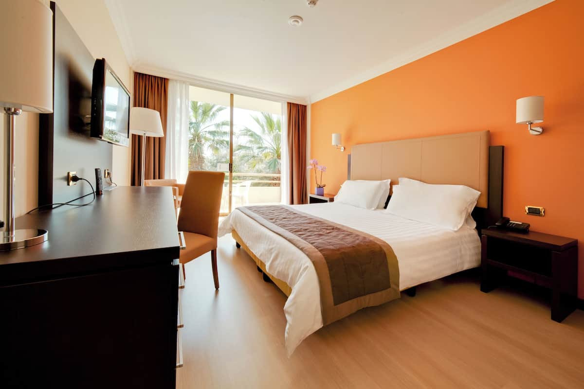 Ein Hotelzimmer mit Bett, Schreibtisch, Stuhl, Fernseher und einem Fenster mit Blick auf Palmen.
