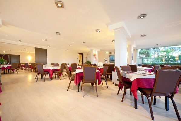 A spacious dining area with multiple tables and chairs, well-lit with ceiling lights, and large windows offering a view of greenery outside. - Nyala Suite Hotel Sanremo (Sanremo)